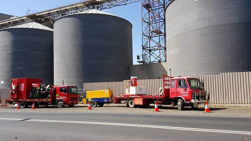 A row of trucks are parked in front of large silos