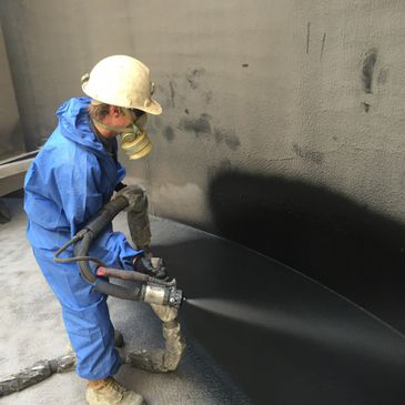 A man in a blue jumpsuit is spraying a black substance on a wall.