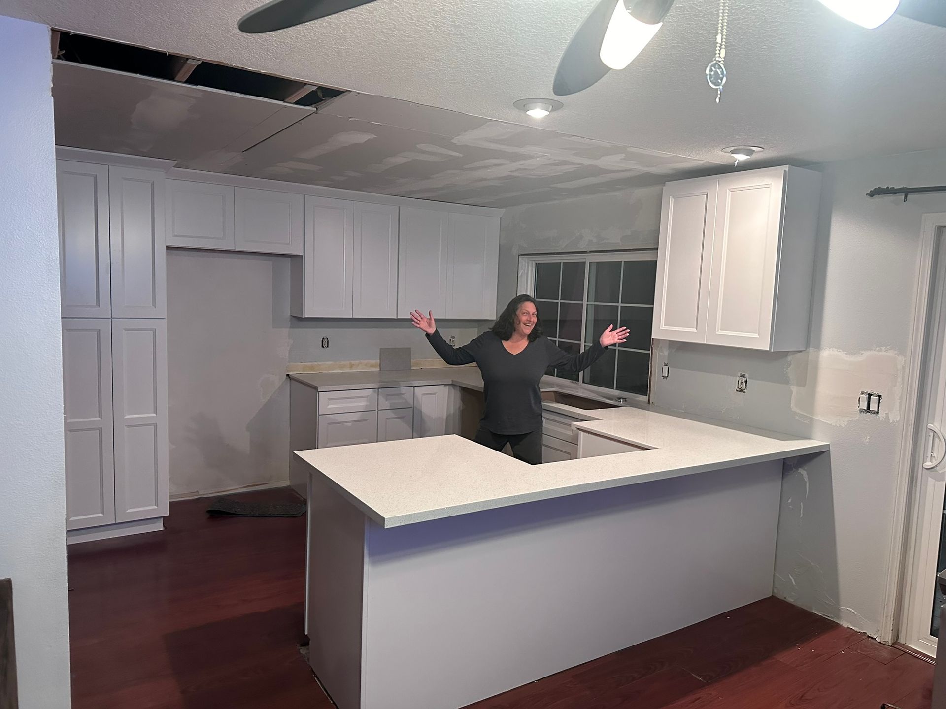 A woman is standing in a kitchen with white cabinets and a ceiling fan.