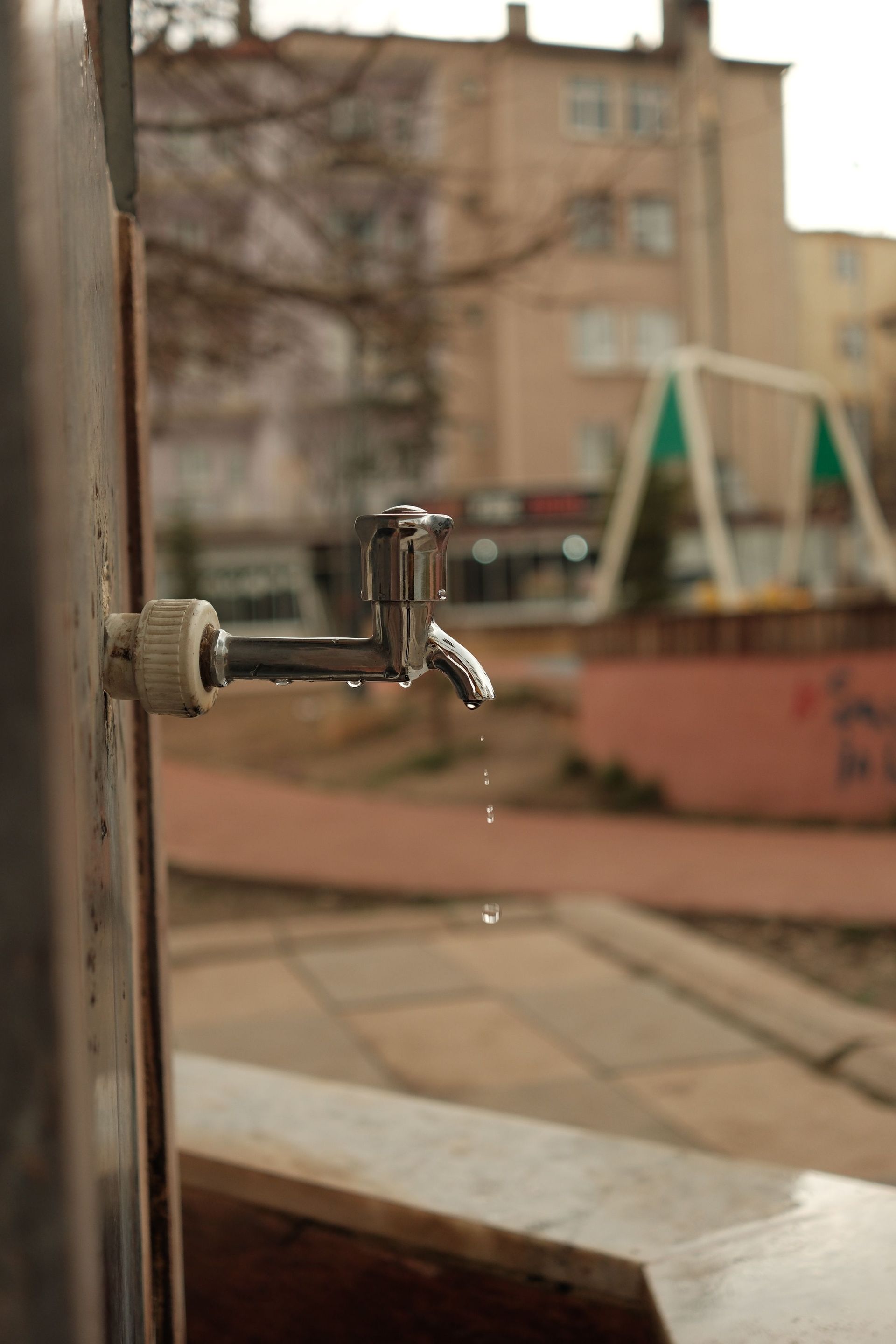 Close-up of a metal faucet on a windowsill with a blurred courtyard and apartment building in the background
