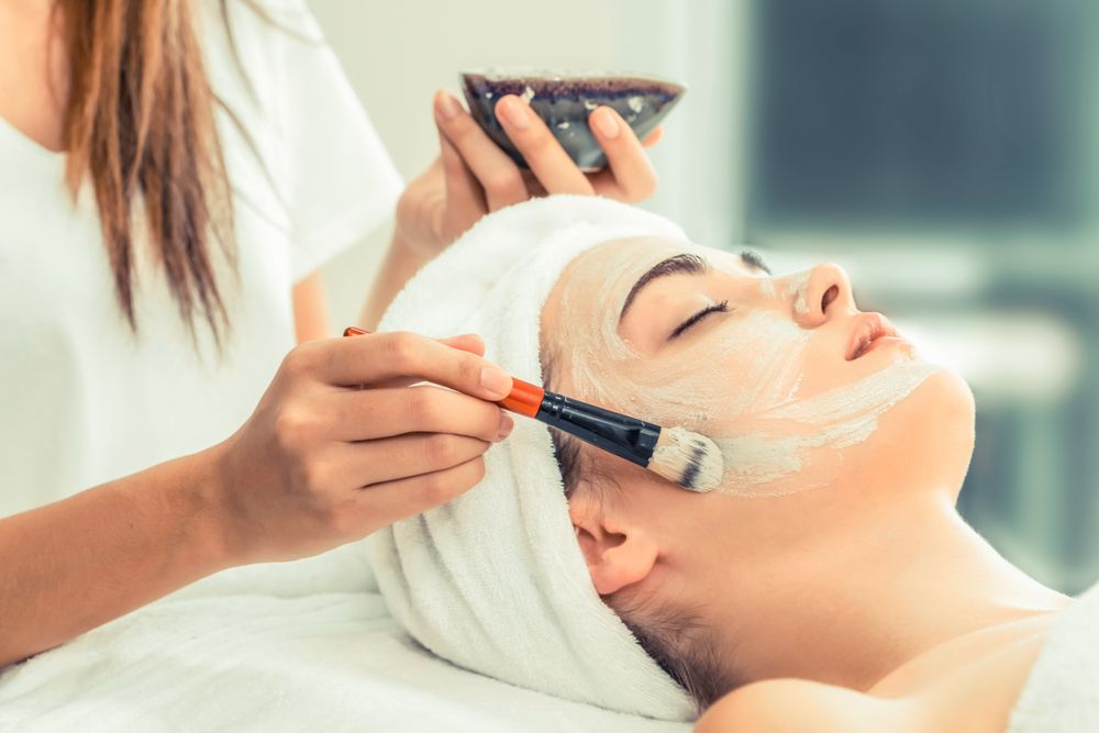 Spa Facial Treatment with A Brush Applying Cream to A Client’s Face While She Reclines with A Towel Wrap — Hope Laser and Beauty Clinic In Forster, NSW