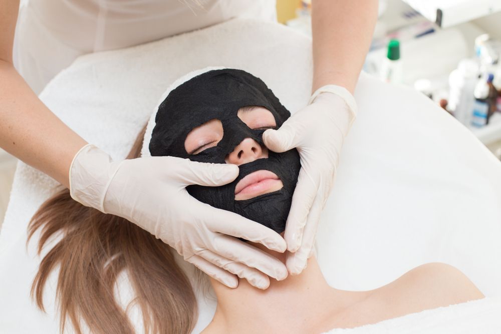 Person Receiving a Black Facial Mask Treatment While Lying Down, with Gloved Hands Adjusting the Mask — Hope Laser and Beauty Clinic In Forster, NSW