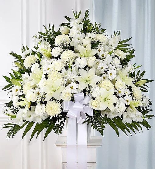 Arreglo floral blanco sobre un pedestal, con lirios y crisantemos, adornado con un lazo blanco.