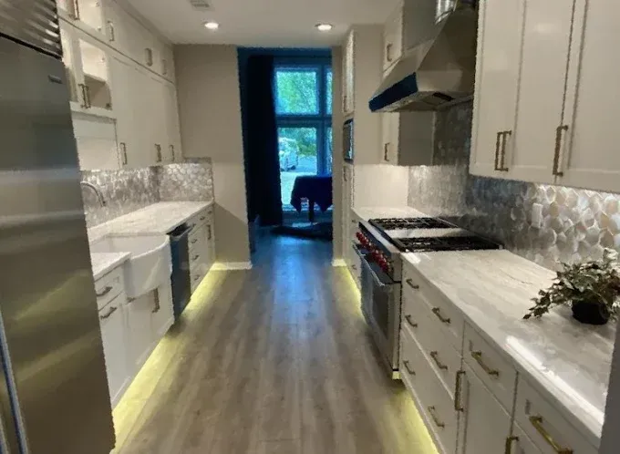 Long, narrow, modern kitchen with white cabinets, marble countertops, stainless steel appliances, and wood flooring.