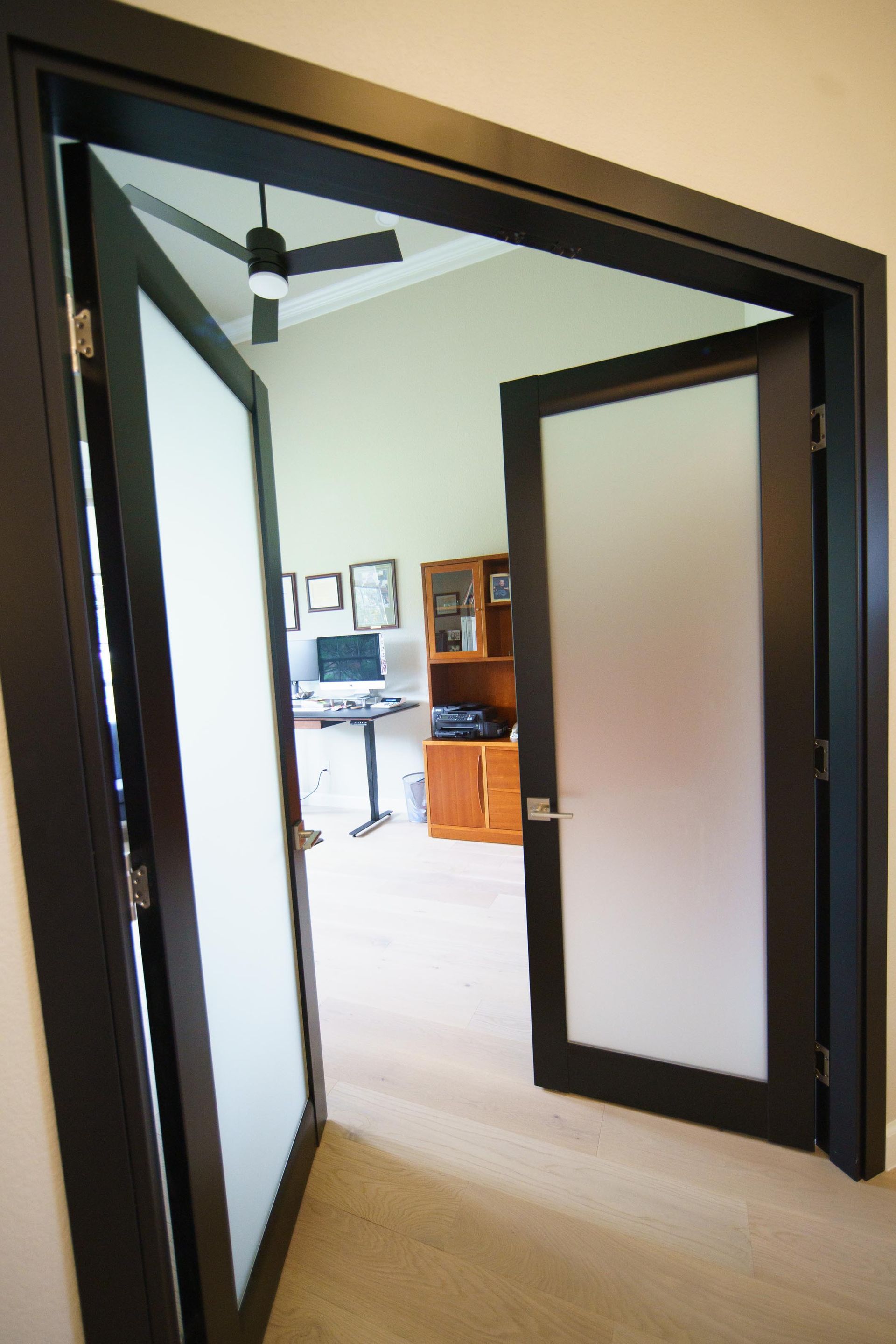 Open, black-framed French doors with frosted glass leading to an office with a desk and wooden cabinet.