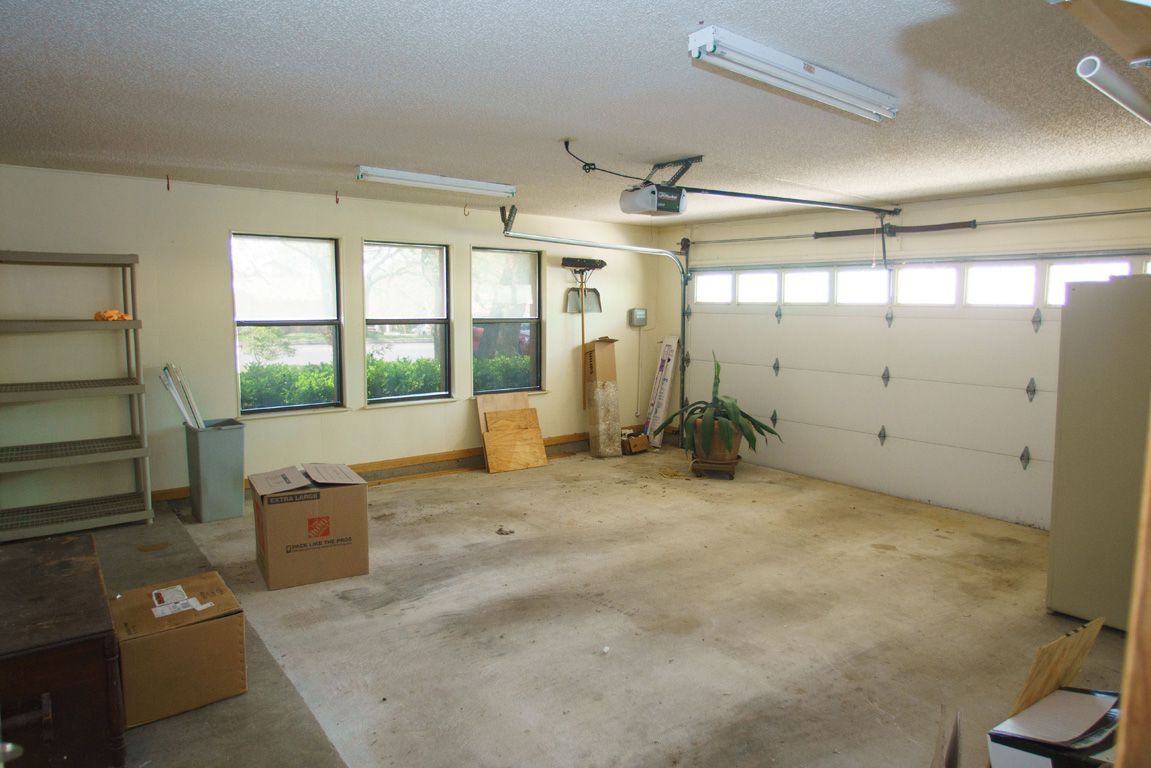 Garage interior with concrete floor, windows, and closed white garage door. Cardboard boxes and shelves present.