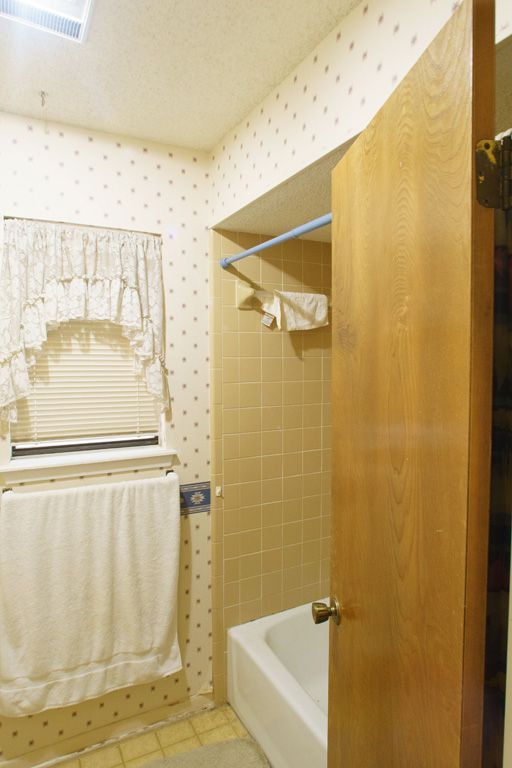 Bathroom interior with light-colored walls and beige tile, featuring a bathtub, window with blinds, and partially opened wooden door.