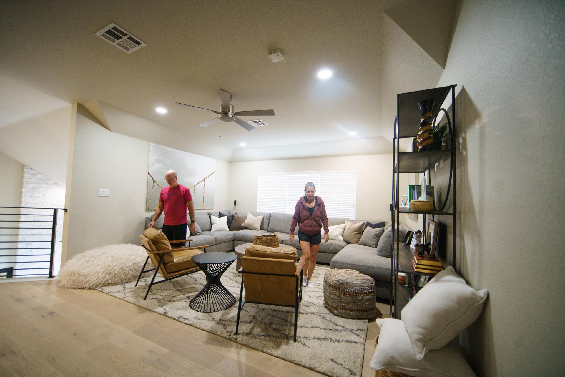Living room with two people near a sectional sofa, rug, and shelving unit.