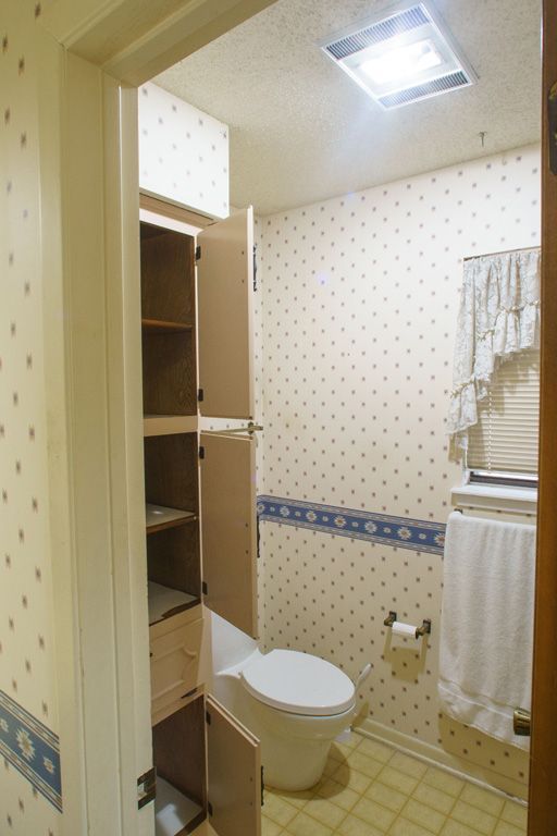 Bathroom with tall storage cabinet, toilet, patterned wallpaper, and window with curtain.