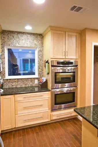Kitchen corner with stacked ovens, light wood cabinets, and mosaic tile backsplash.