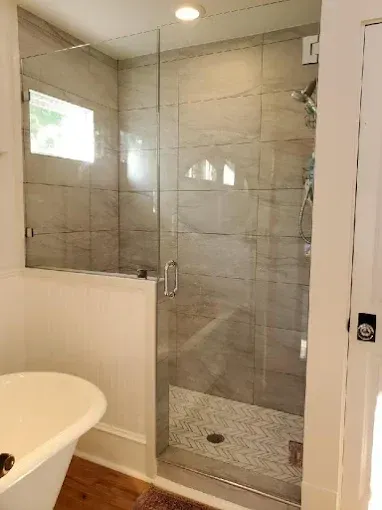 Shower with glass door and gray tiled walls, next to a white tub and a window.