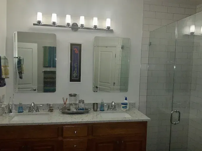 Bathroom with double sinks, mirrors, and a glass shower. White walls, wood cabinets, and bright lighting.