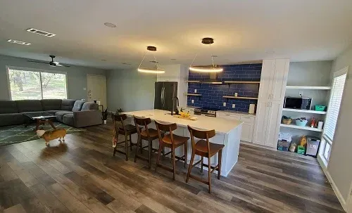 Open-concept kitchen and living area with gray flooring, an island with bar stools, and a blue tile backsplash.