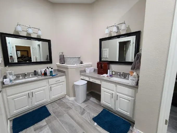 Dual-sink vanity with mirrors, lights, and white cabinets. Blue mats and gray tile floors in a bathroom.