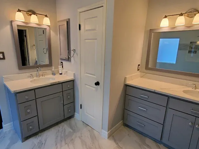Two gray bathroom vanities with mirrors, lights, and a door on a marble floor.