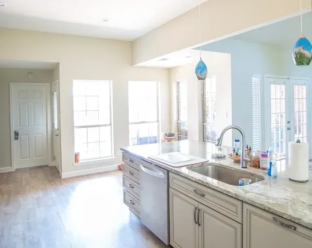 Kitchen with island, sink, cabinets, windows, and door, in a bright, neutral-colored room.