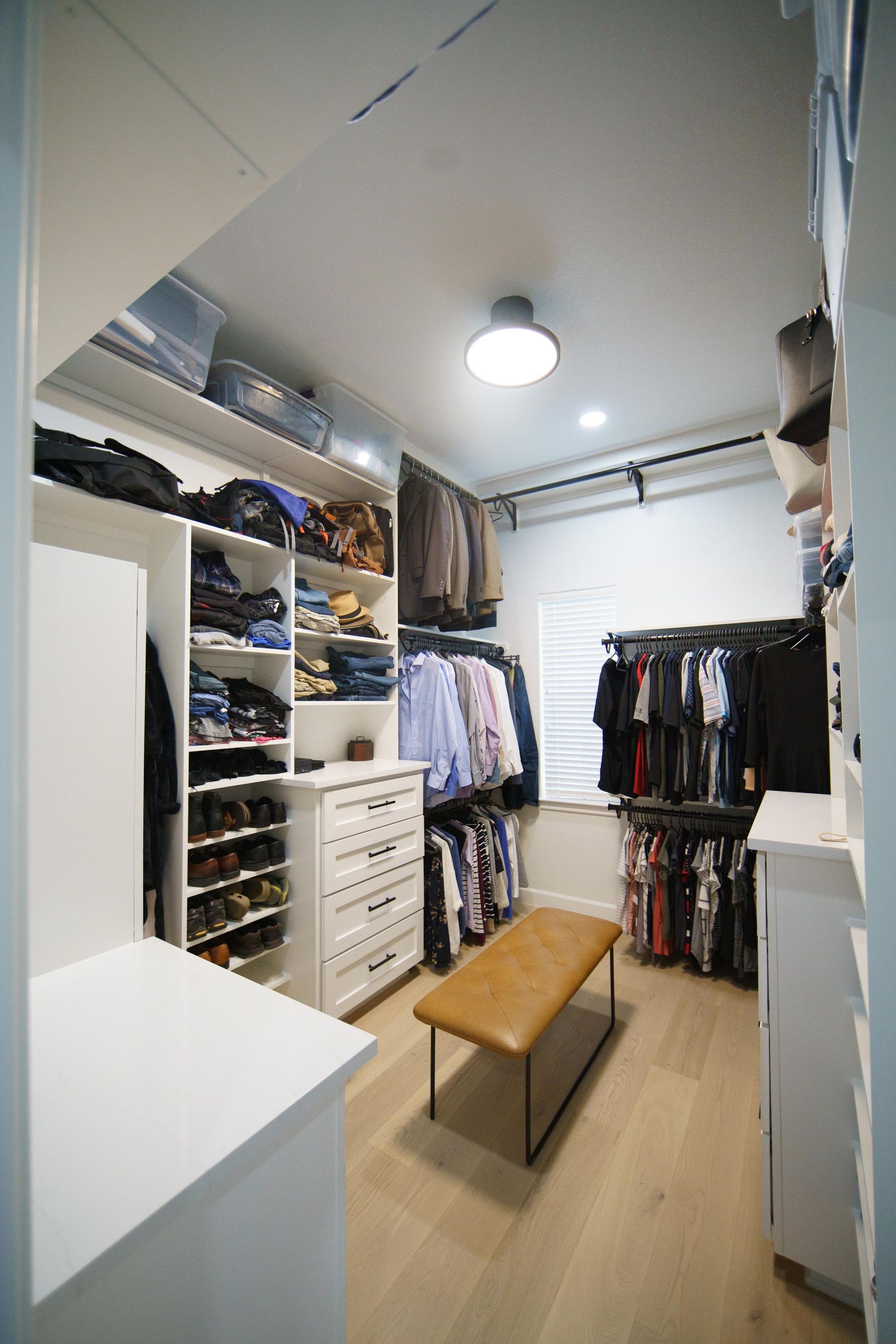 Walk-in closet with white shelves, drawers, and hanging rods filled with clothes. Features a bench and light wood flooring.