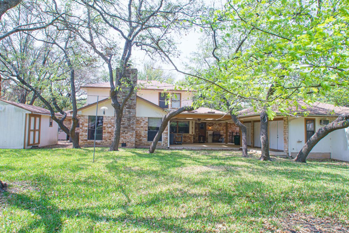 Backyard of a two-story house with a chimney and patio, surrounded by trees and green grass.