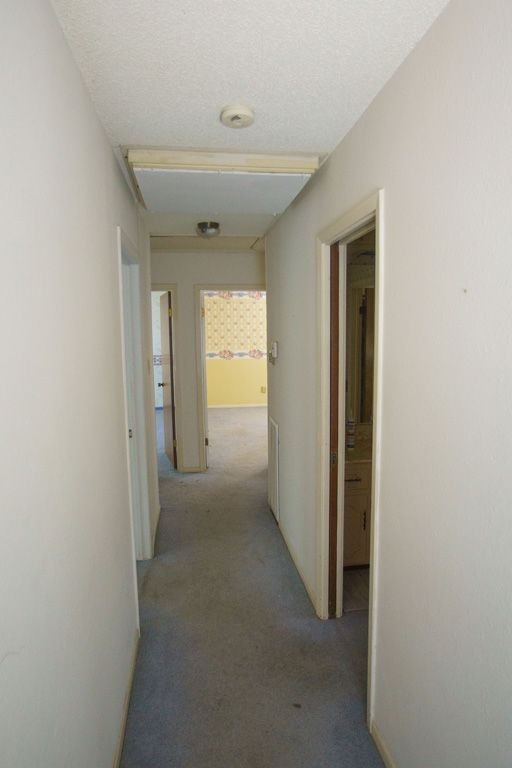 Narrow hallway with doors on either side, leading to a room with yellow wallpaper; gray carpet.