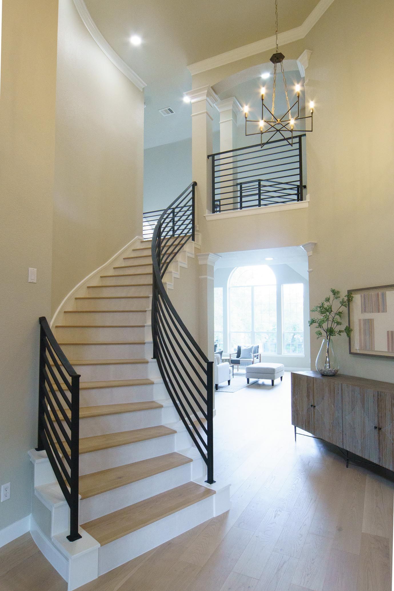 Curving staircase in a light-filled entry with black metal railing. A chandelier hangs above.