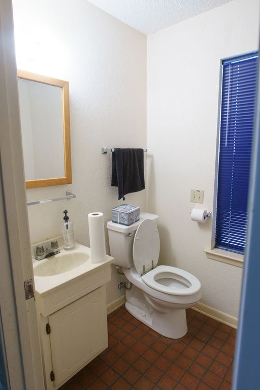 Small bathroom with toilet, sink, mirror, towel bar, and window with blue blinds. Red-brown tiled floor.