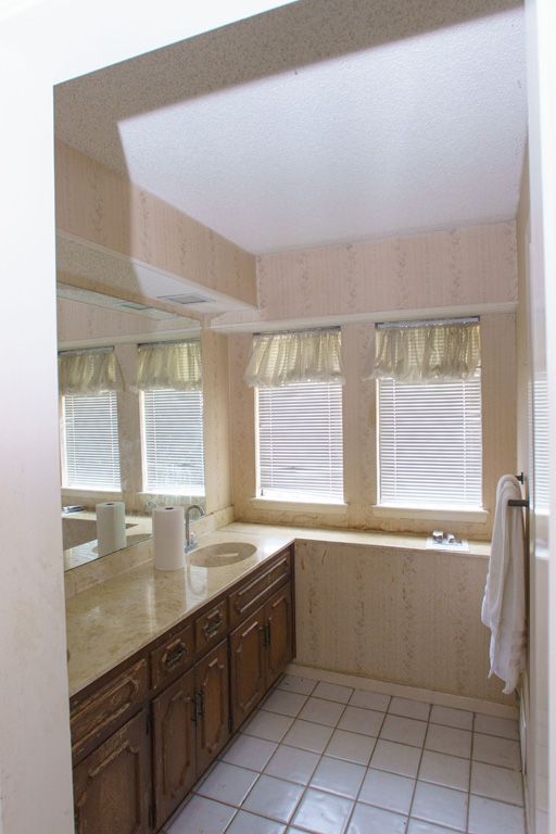 Bathroom with beige walls, marble counter, wooden cabinets, three windows with blinds, and white tile floor.