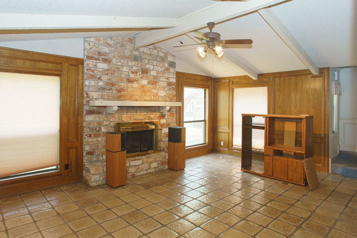 Empty living room with brick fireplace, wood paneling, tile floor, and large windows.