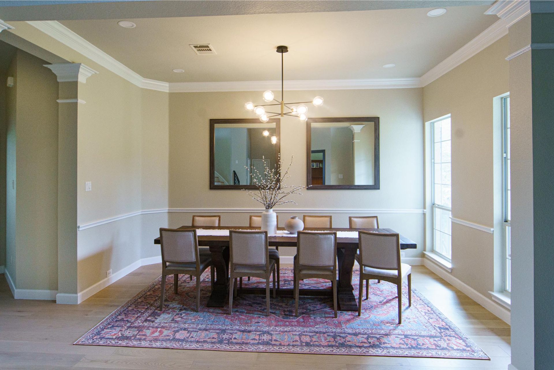 Dining room with long wooden table, eight chairs, two mirrors, and a floral rug.