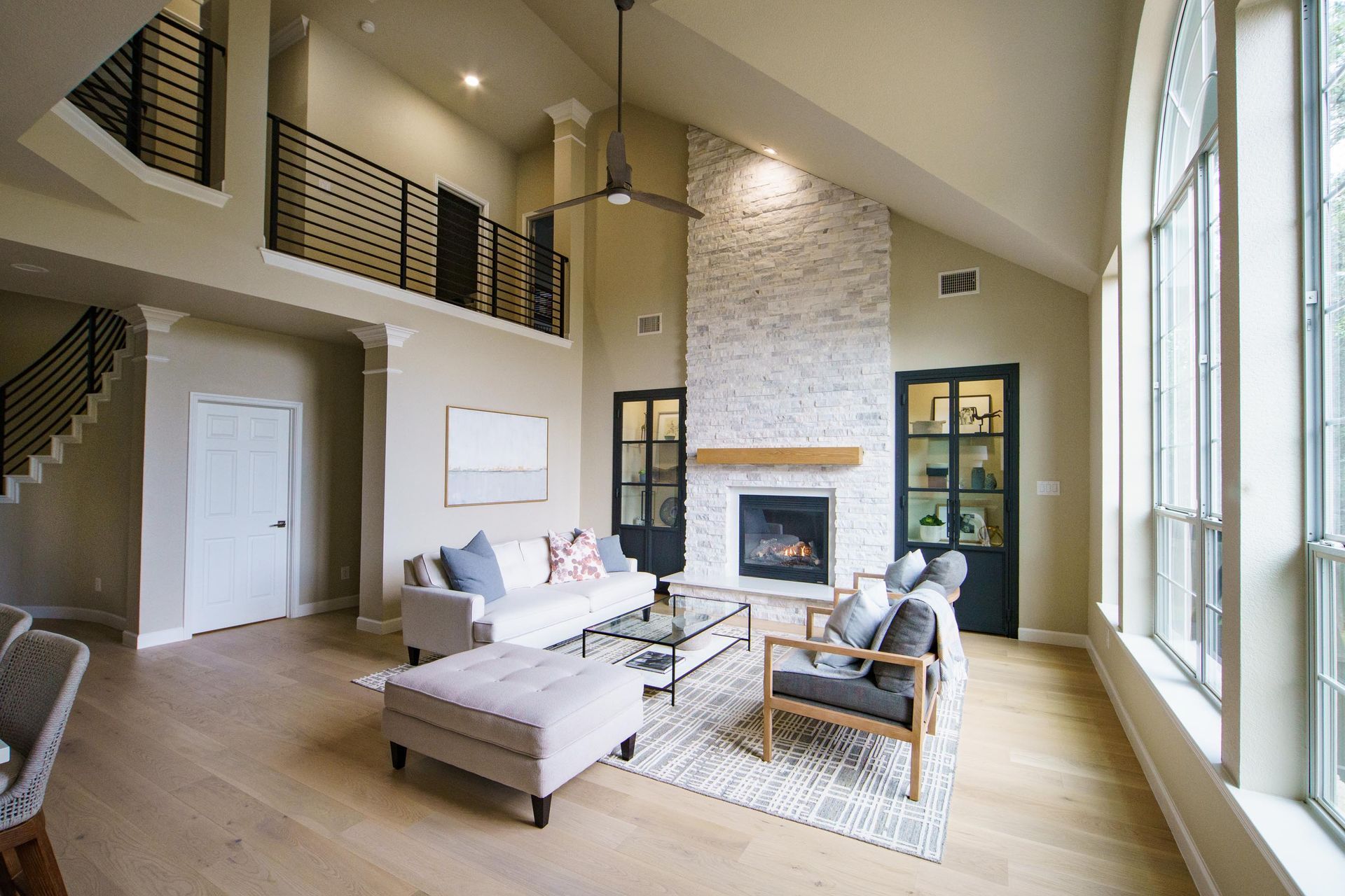 Living room with high ceiling, brick fireplace, hardwood floors, and large windows.