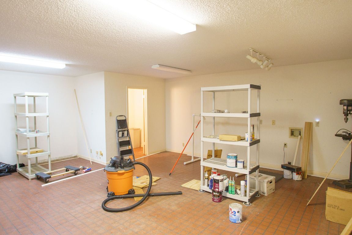 A cluttered, empty room with shelving, a vacuum, and various tools, likely a workshop or storage area.