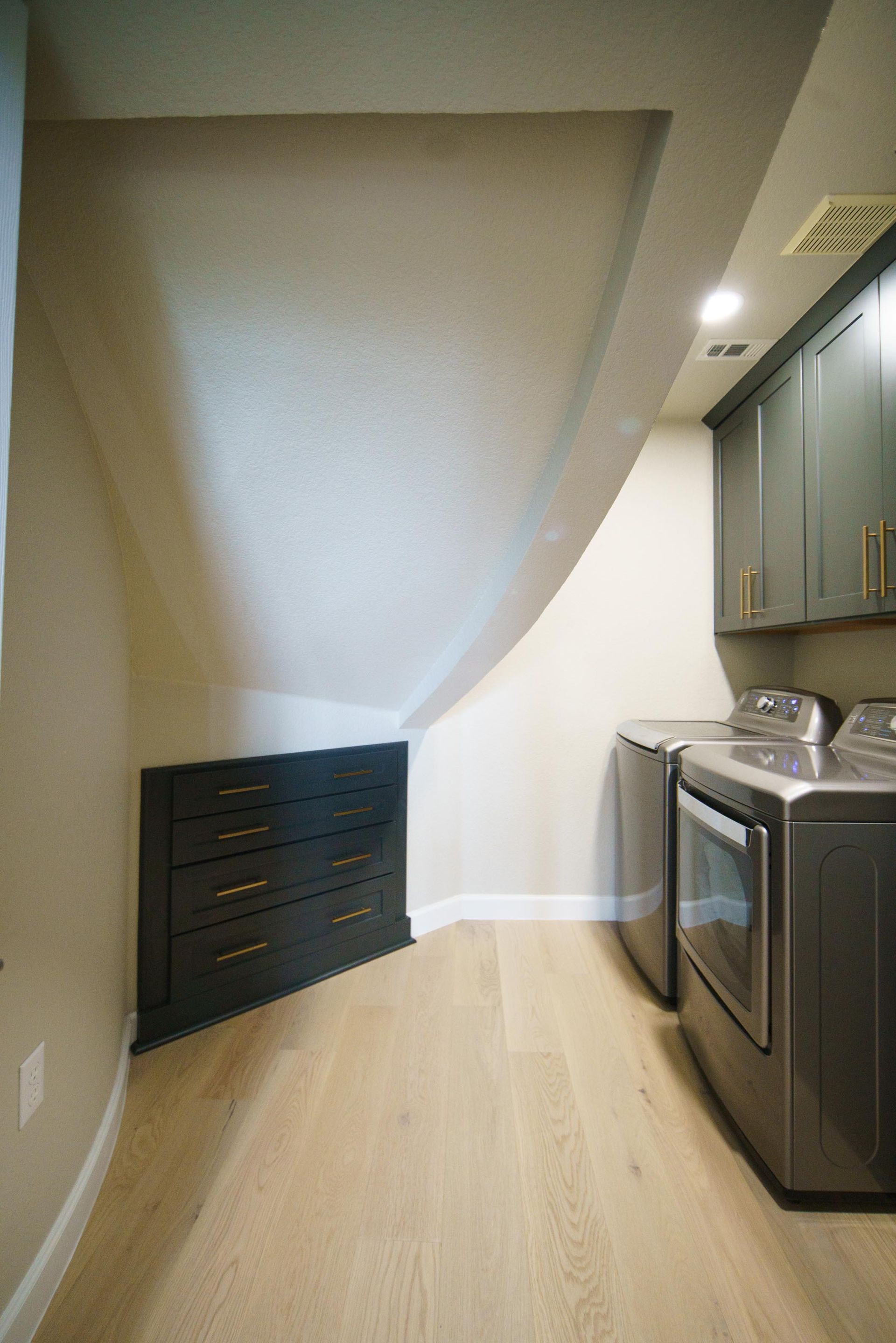 Laundry room with slanted ceiling, gray washer and dryer, dark drawers, and gray cabinets.