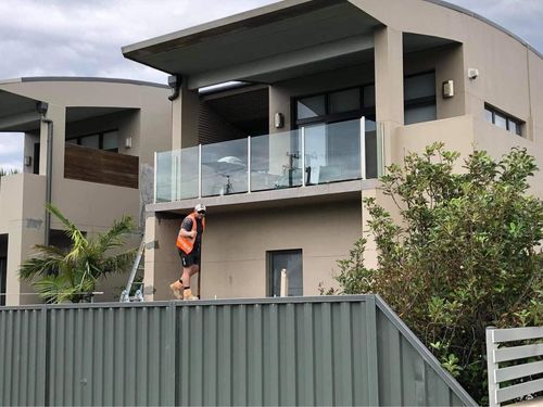 Worker standing on a ladder beside a modern rendered house with a glass balcony and green fence
