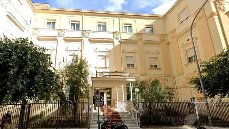 Uomo in camice verde acqua che scrive alla scrivania, con occhiali e orologio. Ambientazione interna, con documenti.