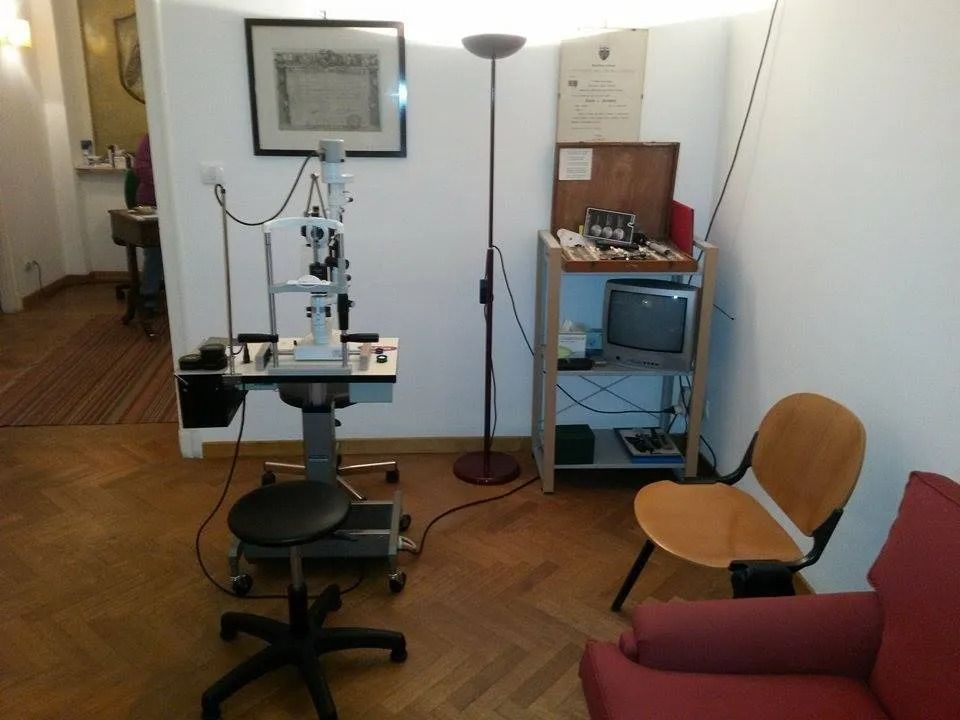 Sala per visita oculistica con lampada a fessura, sedia, scaffale per le attrezzature e poltrona rossa.