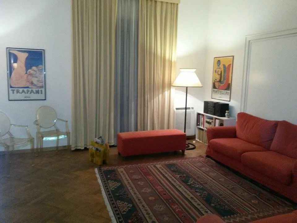 Soggiorno con divano rosso, tappeto, pouf e tende; opere d'arte alle pareti.