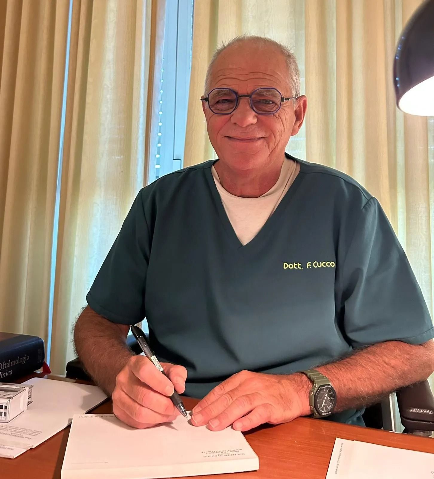 Uomo con gli occhiali, che scrive alla scrivania, indossa un camice verde acqua, guarda la telecamera e sorride.