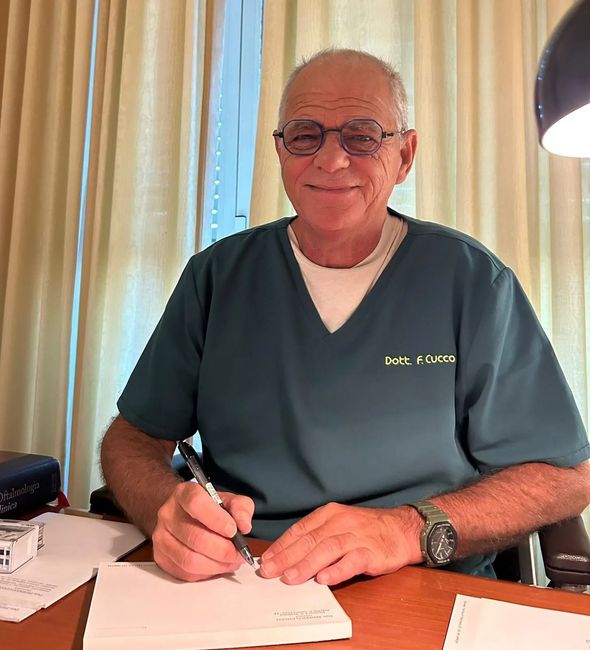 Uomo con gli occhiali, che scrive alla scrivania, indossa un camice verde acqua, guarda la telecamera e sorride.