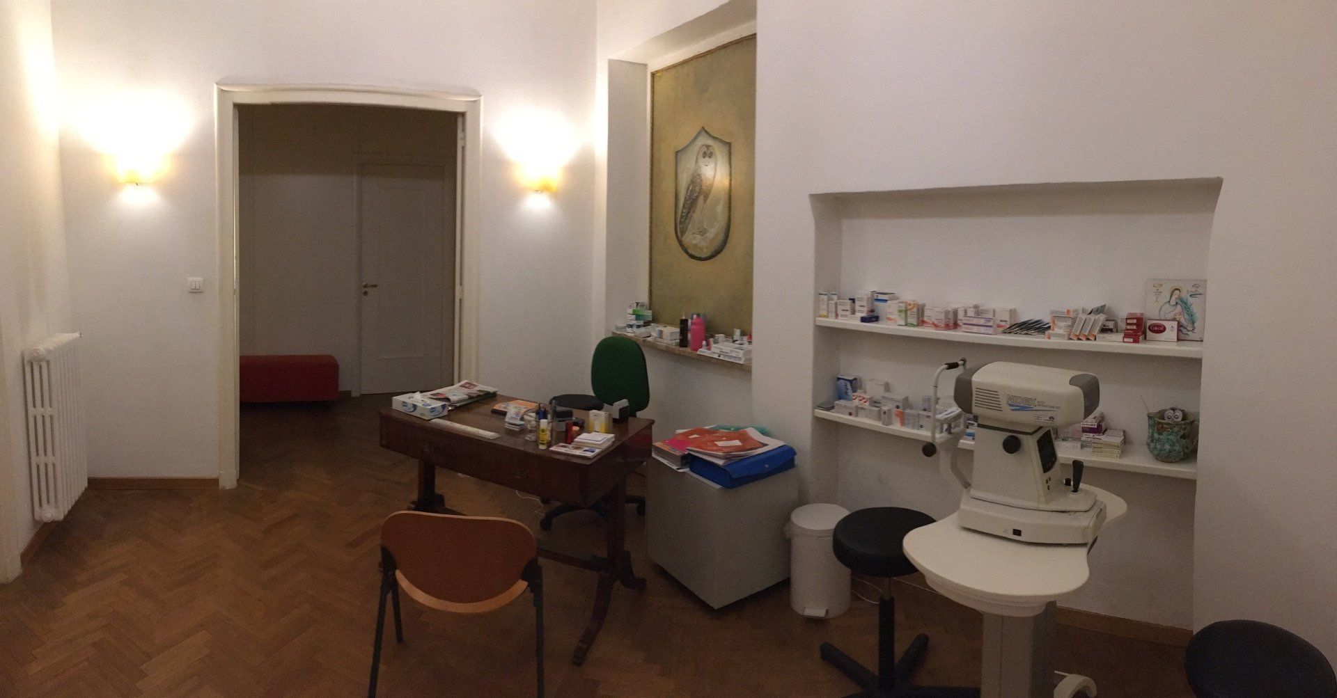 Interno di una stanza con scrivania, robot, scaffali e porta aperta. Pareti bianche, pavimento in legno, luci soffuse.