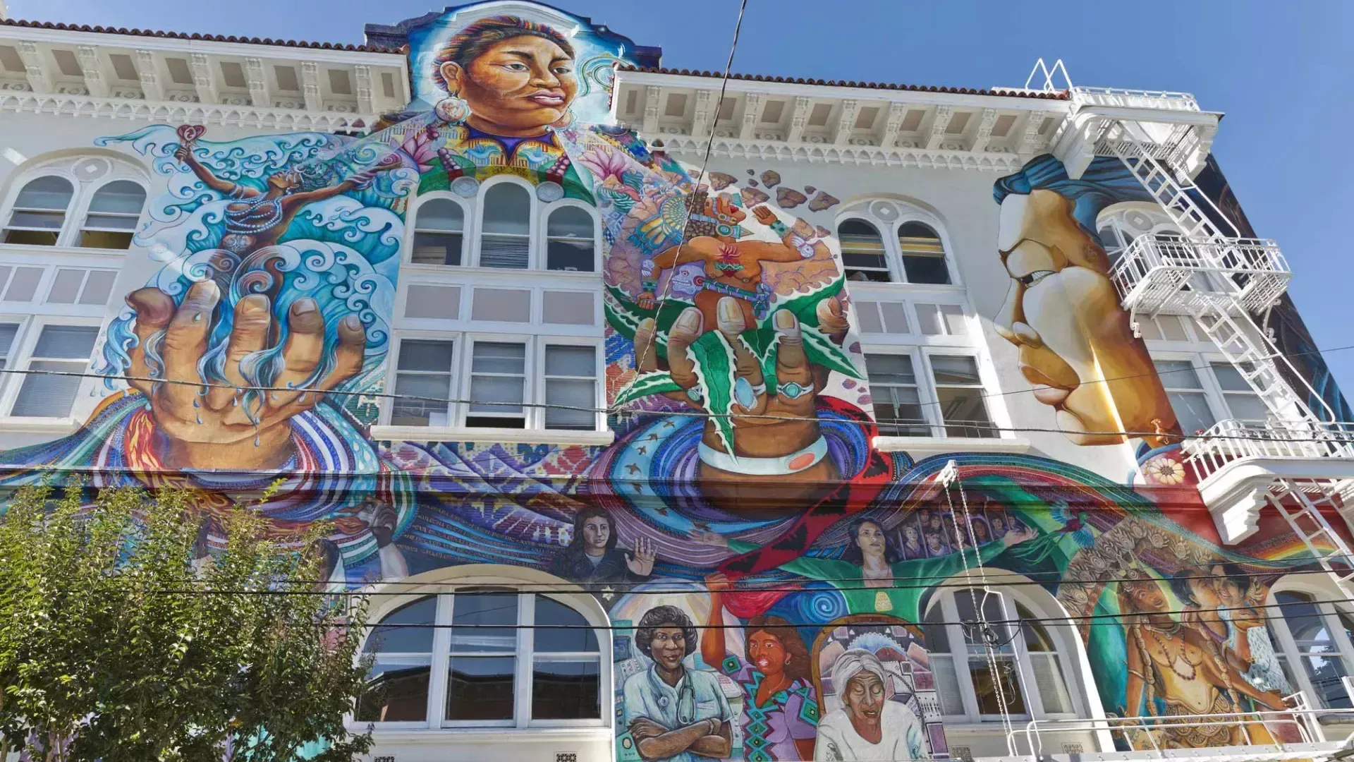 A colorful, large-scale mural covers the entire facade of a white building, featuring figures, hands, and intricate patterns.