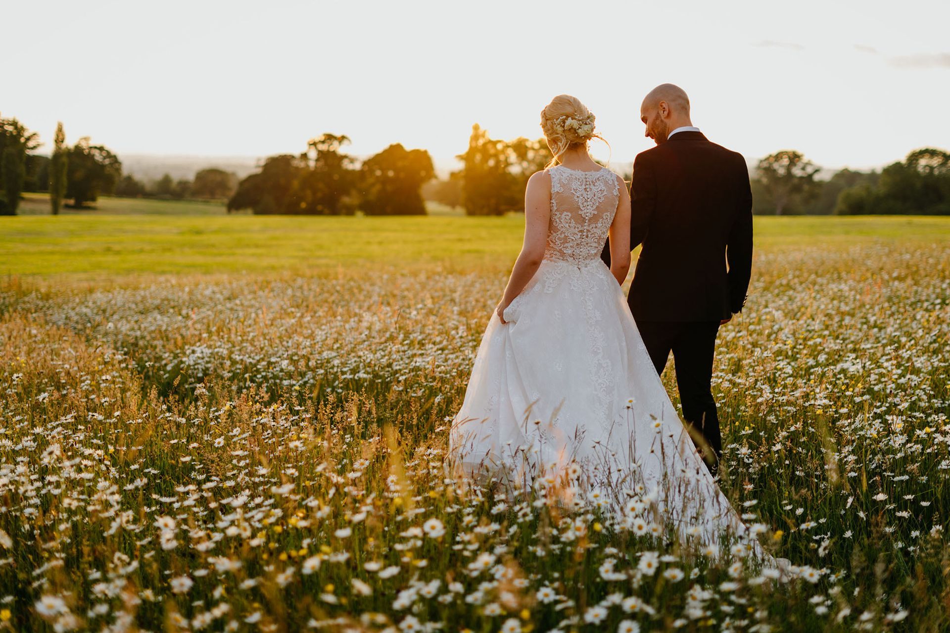 a bride and groom are walking through a field of daisies at sunset .