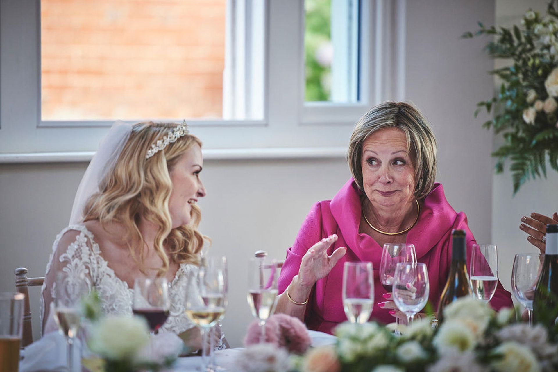 a bride and her mother are sitting at a table with wine glasses.