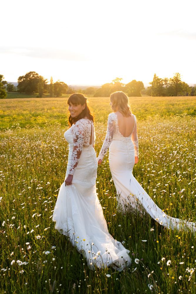 two women in wedding dresses are walking through a field of flowers .