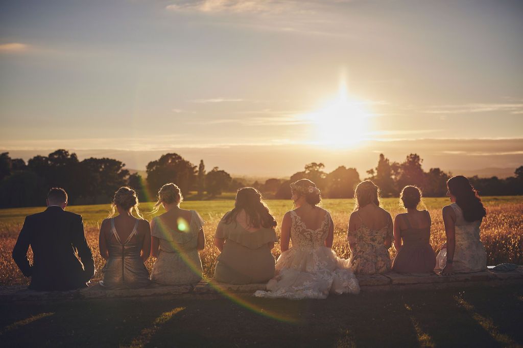a bride and groom and their wedding party are sitting in a field at sunset .