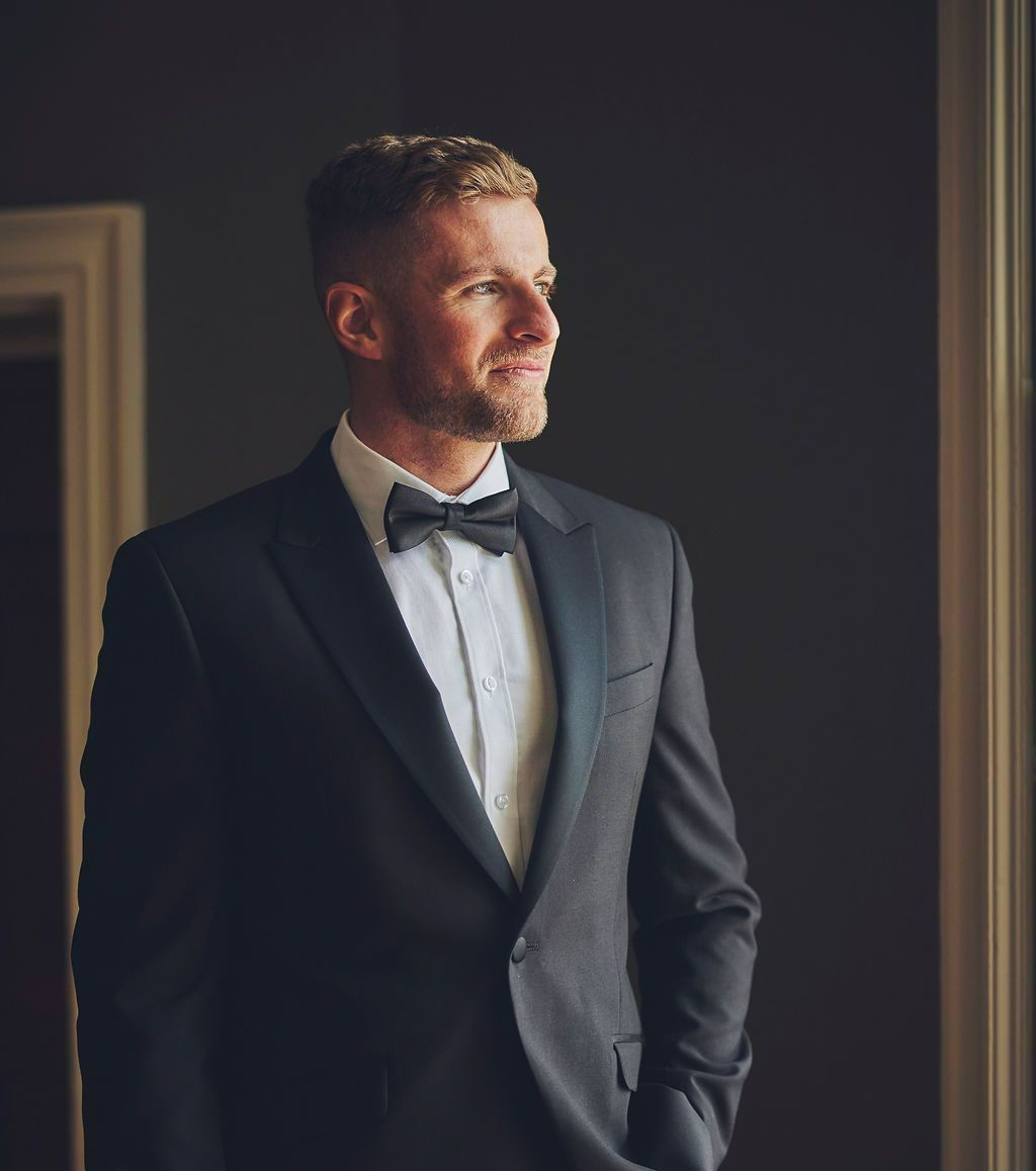 a man in a suit and bow tie is standing in front of a window .