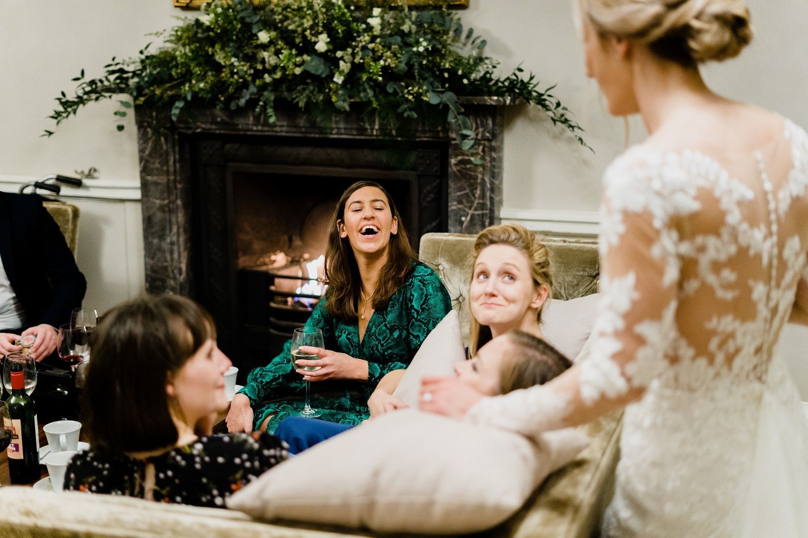 a bride is standing next to a group of women sitting on a couch in front of a fireplace .