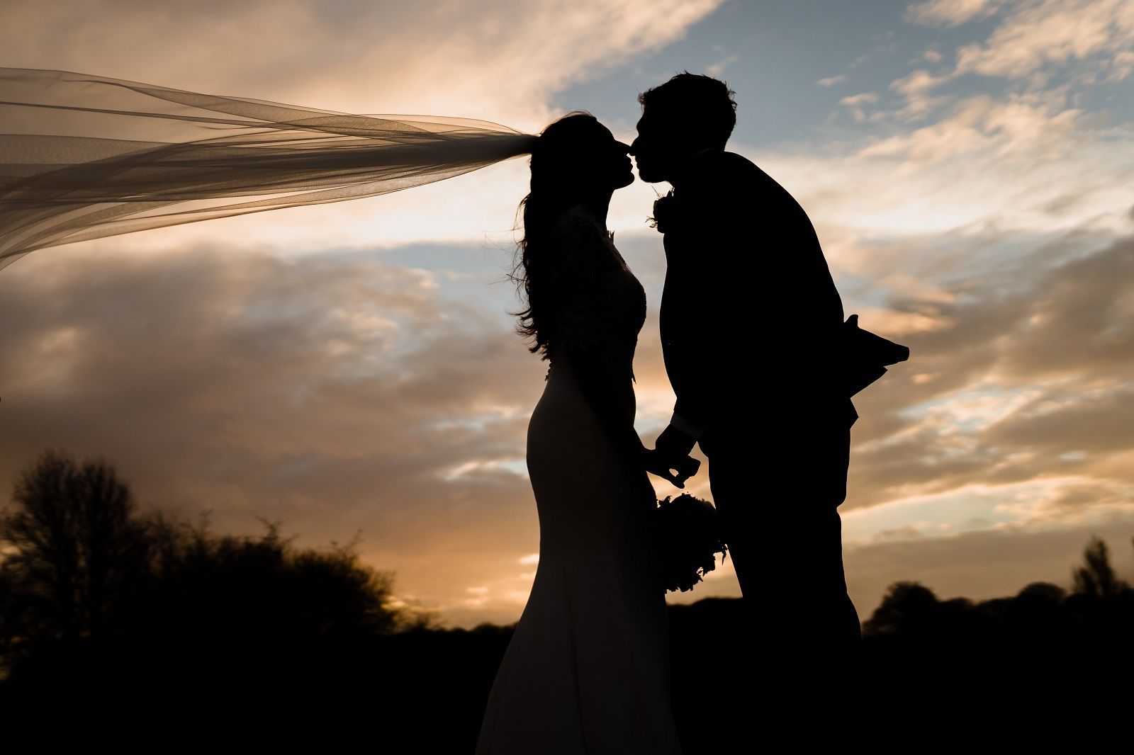 a bride and groom are kissing at sunset with their veil blowing in the wind .