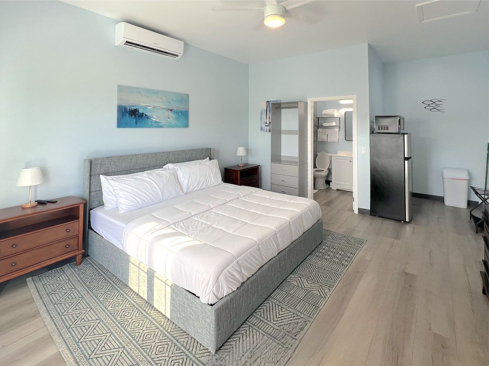 Bedroom with a gray bed, light blue walls, a rug, and a fridge. The bathroom is visible in the background.