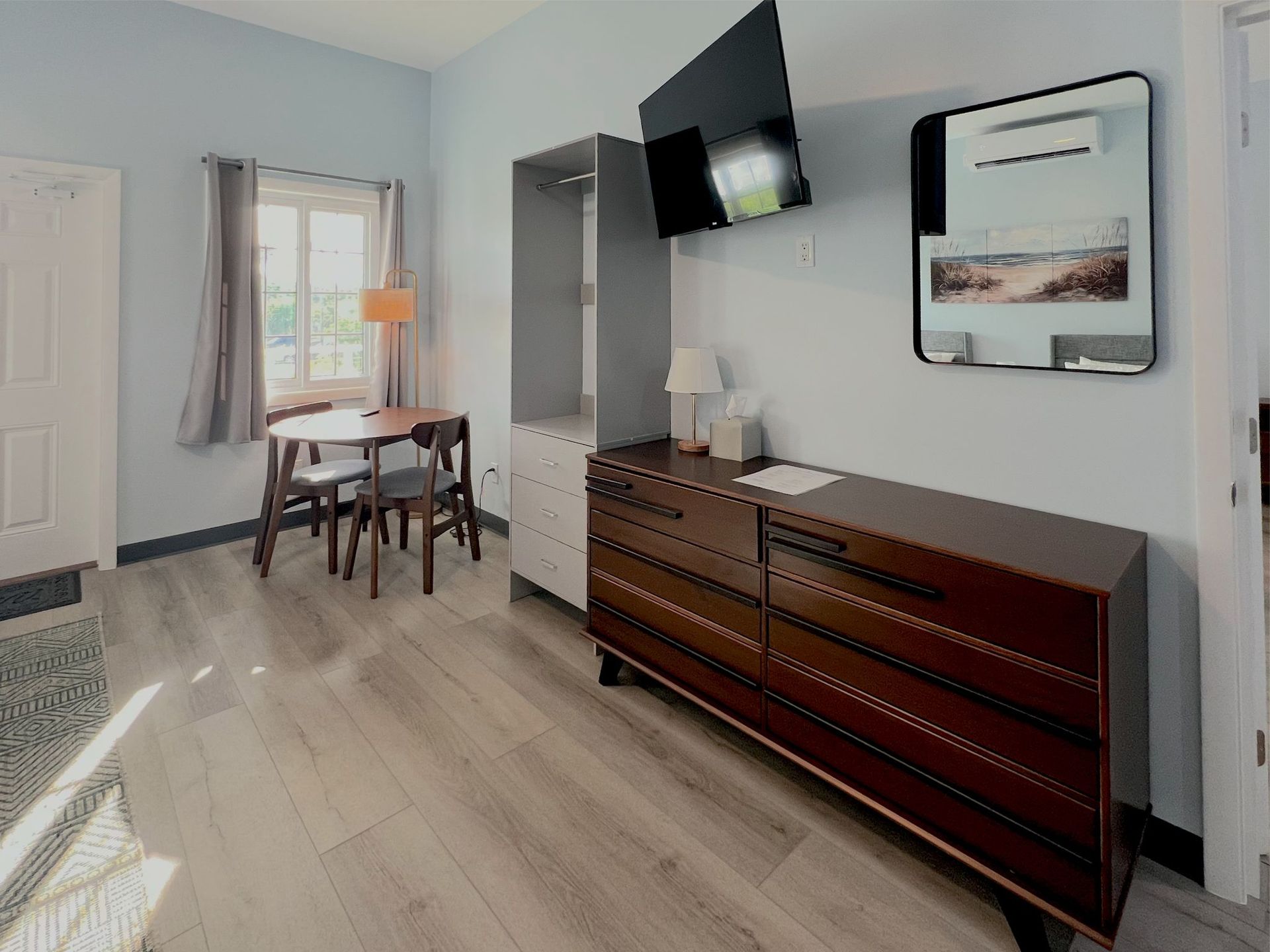 Small room with wooden furniture, including a dresser, table, and tall cabinet; a TV and mirror are mounted on the wall.