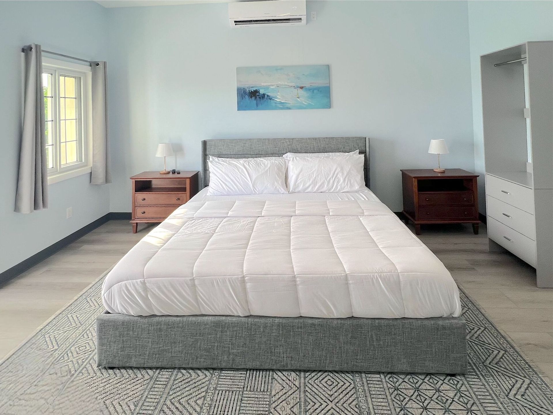 Bedroom with bed, nightstands, rug, wardrobe, window. Light blue walls, white bedding, wood furniture.