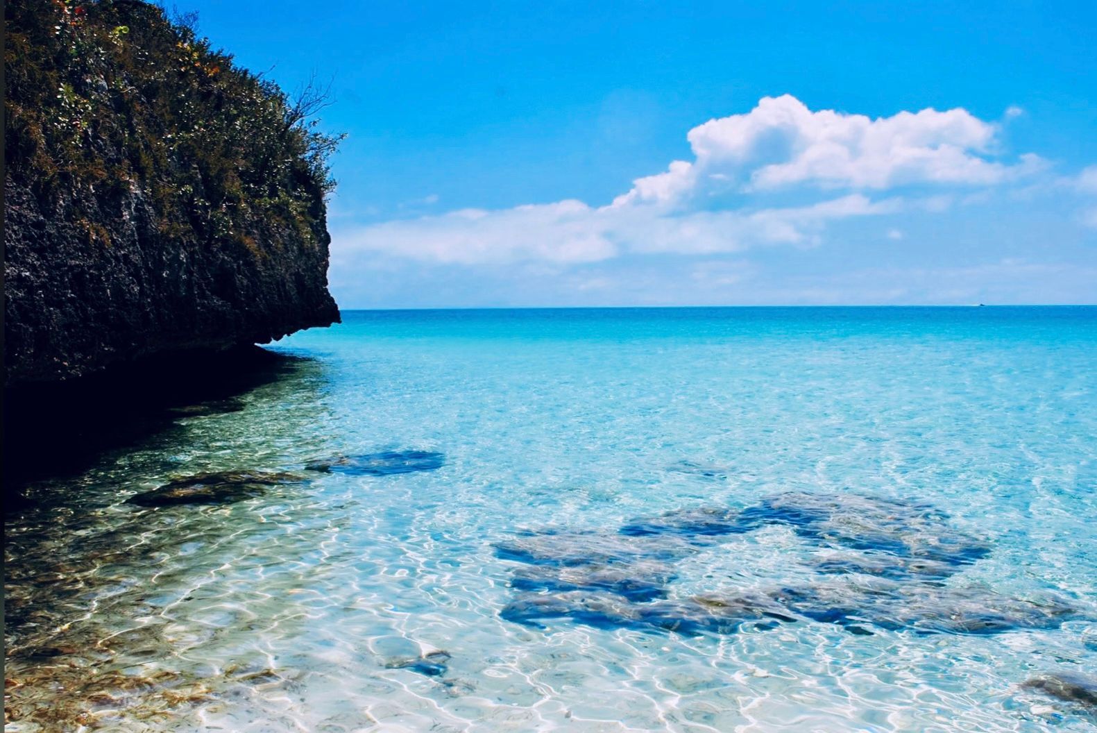 Clear turquoise water, a rocky cliff with sparse greenery, and a bright blue sky with puffy white clouds.